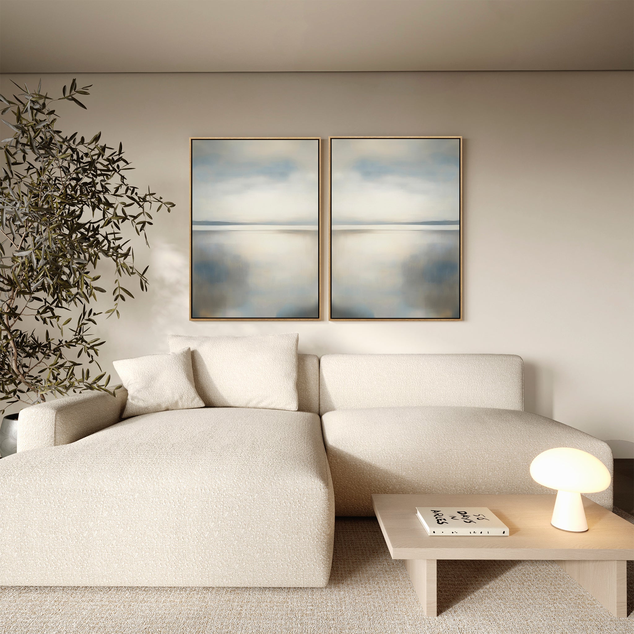 A minimalist living room features a cream sectional, light wood coffee table, tall potted plant, and the Still Horizon Set of 2 wall art in abstract blue and beige, evoking the calm of a serene shoreline.