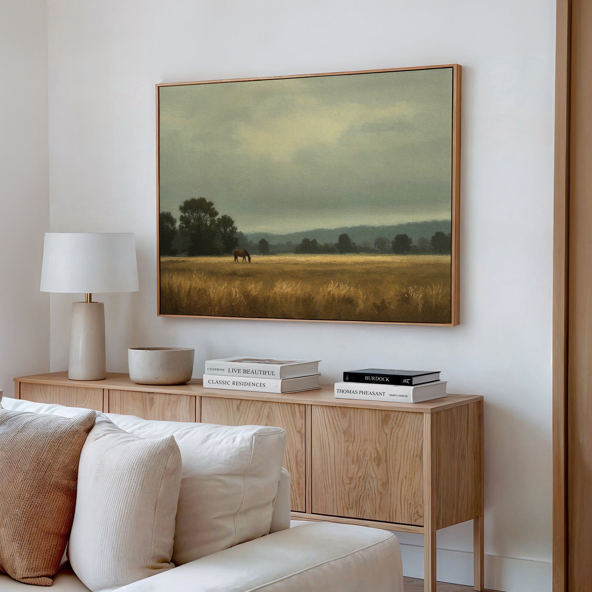 A cozy living room featuring a light wood sideboard, white lamp, stacked books, bowl, and countryside wall art—the Still Morning Canvas Art depicting a misty field with a horse and trees—hanging above.