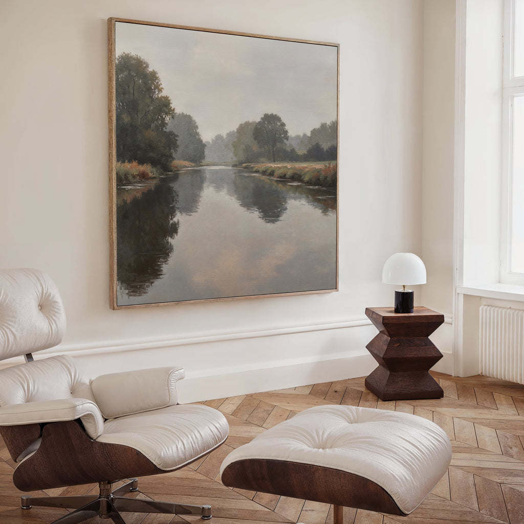 A minimalist room with a leather lounge chair and ottoman, a wooden side table with a white lamp, and the Still Riverbank Canvas Art above. Wooden floors and natural light from a large window complete this tranquil bedroom decor.