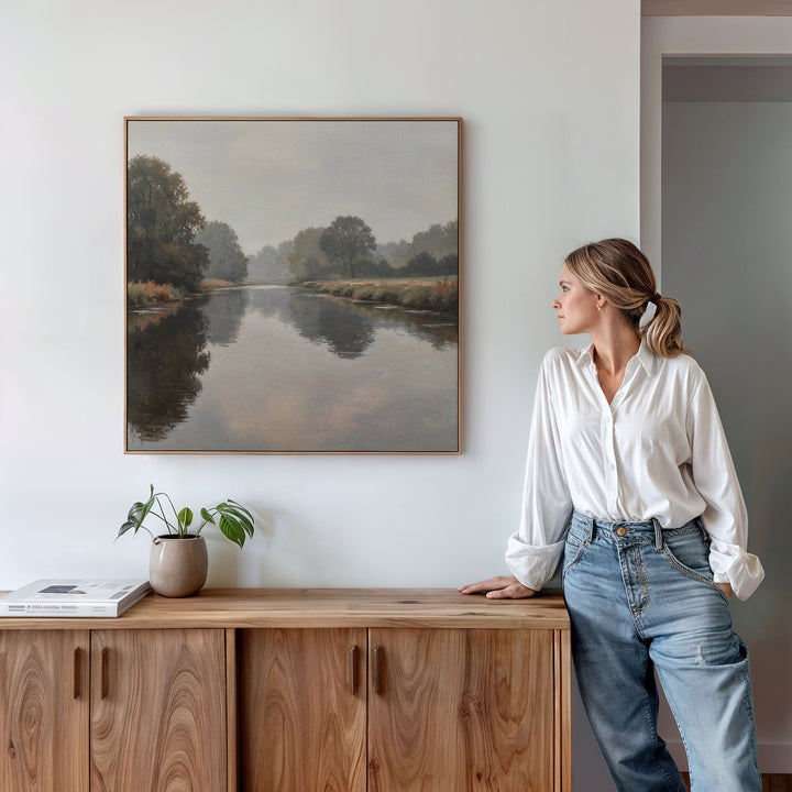 A woman in a white blouse and jeans leans against a wooden cabinet, admiring the Still Riverbank Canvas Art displayed above. A potted plant and books sit on the cabinet, enhancing the tranquil bedroom ambiance.