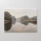 The Still Riverbank Canvas Art – Horizontal features a calm river reflecting trees and an overcast sky in soft, muted colors, ideal as minimalist wall art for serene bedroom decor. Displayed mounted on a white wall.