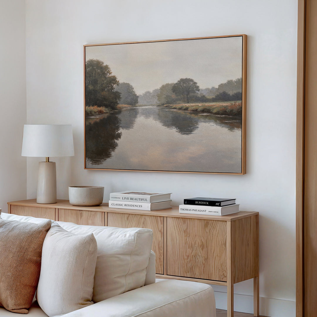 A cozy living room corner features a wooden sideboard, beige lamp, decorative bowl, stacked books, and the Still Riverbank Canvas Art – Horizontal—a minimalist landscape painting of a calm river—adding style to the white wall.