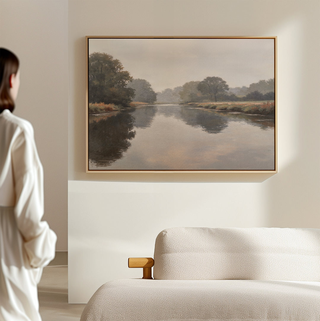 A woman in a white shirt admires the Still Riverbank Canvas Art – Horizontal, a minimalist landscape of a calm river and trees, framed above a beige sofa on a light wall, adding tranquil elegance to the space.