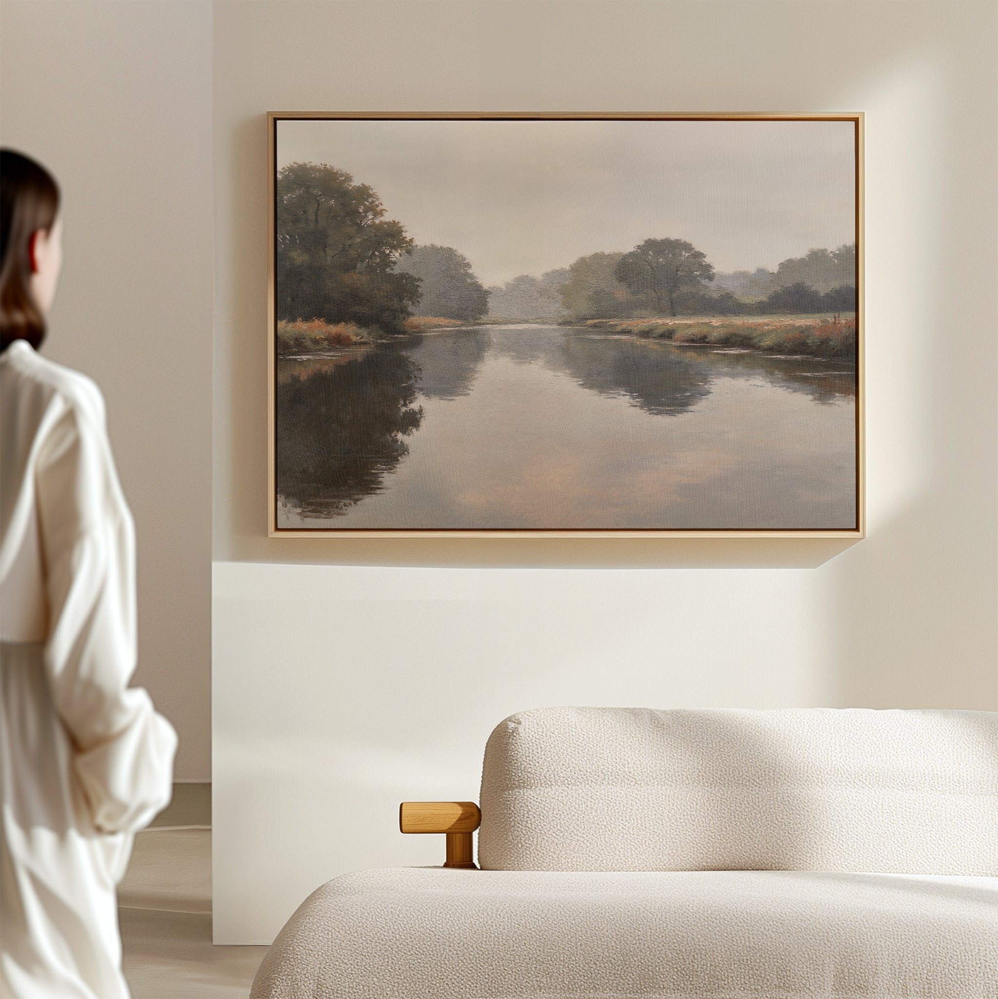 A woman in a white shirt admires the Still Riverbank Canvas Art – Horizontal, a minimalist landscape of a calm river and trees, framed above a beige sofa on a light wall, adding tranquil elegance to the space.