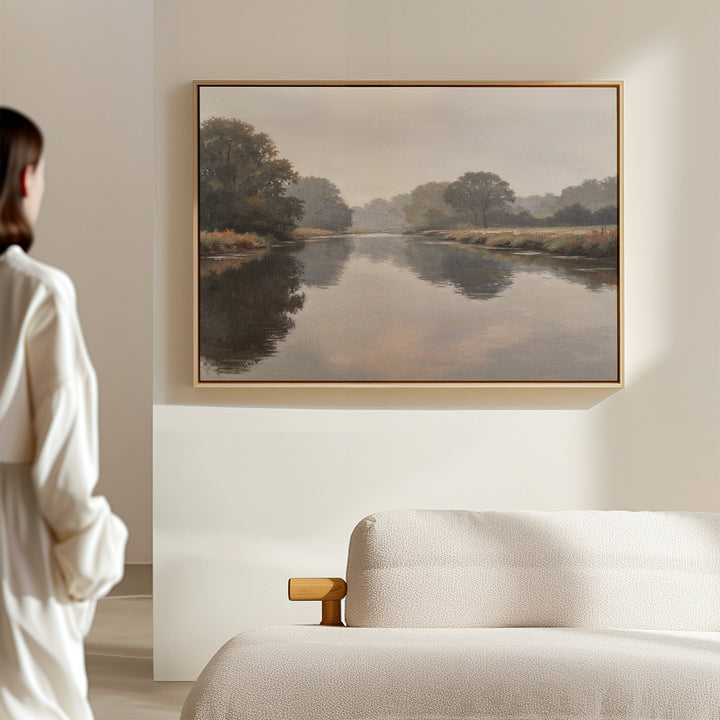 A woman in a white shirt admires the Still Riverbank Canvas Art – Horizontal, a minimalist landscape of a calm river and trees, framed above a beige sofa on a light wall, adding tranquil elegance to the space.