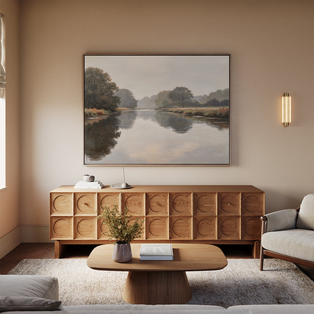 A minimalist living room features a wooden coffee table, textured sideboard, light sofa, and the Still Riverbank Canvas Art – Horizontal above the sideboard. A wall sconce casts warm light, enhancing the serene atmosphere.