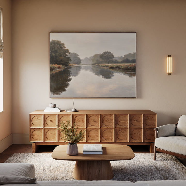A minimalist living room features a wooden coffee table, textured sideboard, light sofa, and the Still Riverbank Canvas Art – Horizontal above the sideboard. A wall sconce casts warm light, enhancing the serene atmosphere.