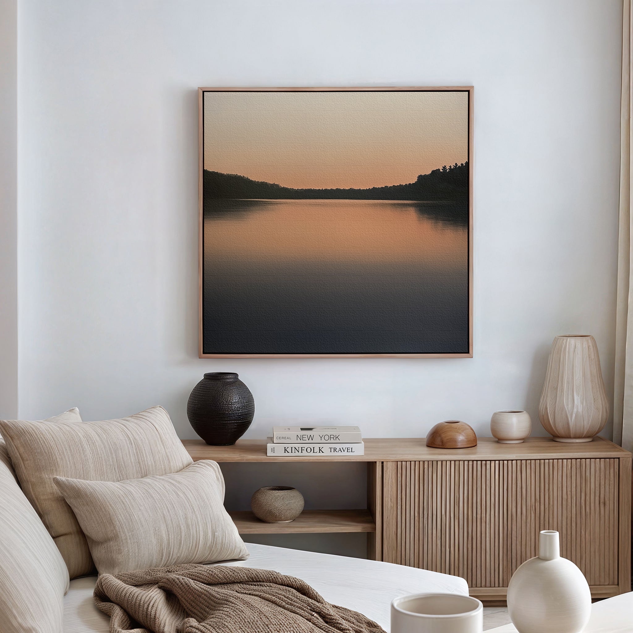 A serene living room with a beige sofa, striped cushions, a wooden sideboard, vases, books, and peaceful decor is enhanced by Still Waters Canvas Art—a large framed landscape painting of a lake at sunset.