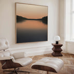 A modern, minimalist living room features a white leather lounge chair and ottoman, a wooden side table with a round white lamp, and the Still Waters Canvas Art—a large tranquil lake sunset print—perfect for stylish decor.