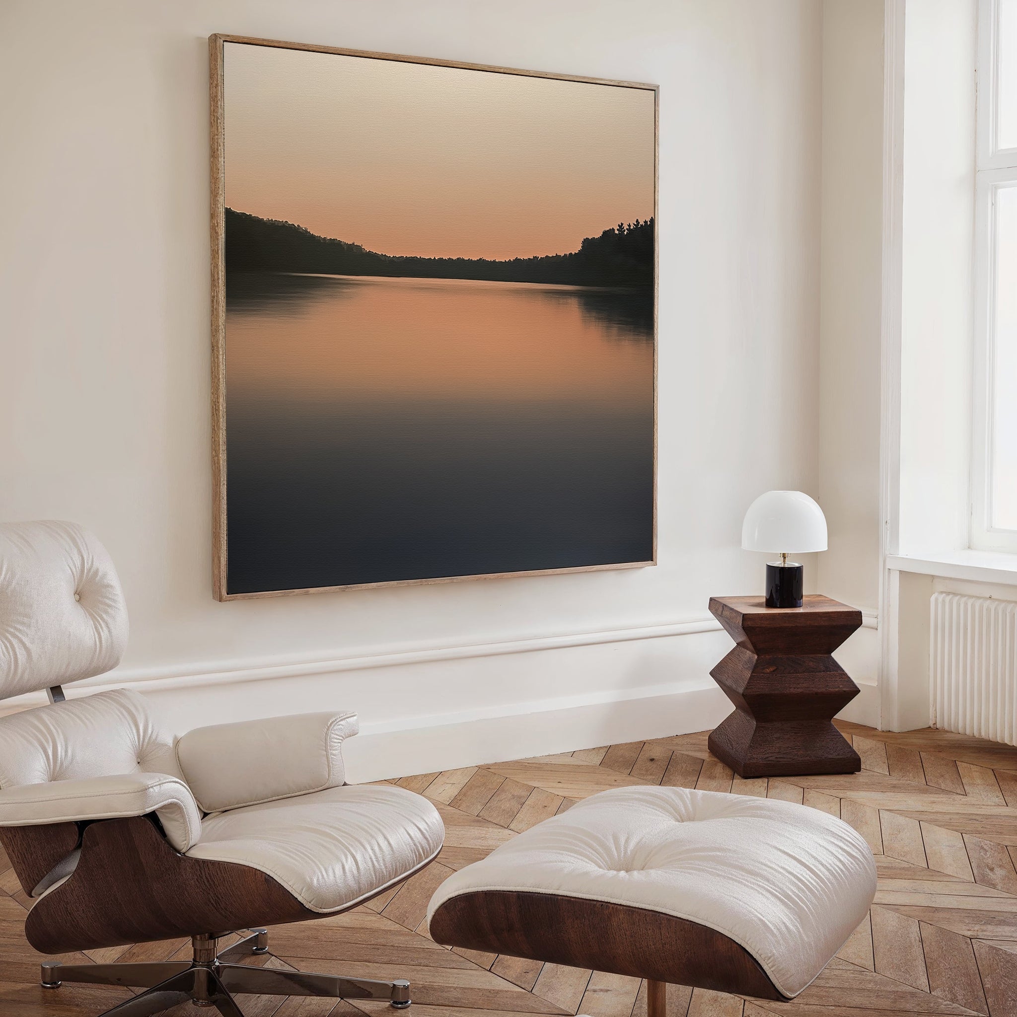 A modern, minimalist living room features a white leather lounge chair and ottoman, a wooden side table with a round white lamp, and the Still Waters Canvas Art—a large tranquil lake sunset print—perfect for stylish decor.