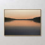 The Still Waters Canvas Art – Horizontal features a tranquil lake at sunset with soft orange and pink hues reflected on the water, dark tree silhouettes lining the horizon—perfect as serene bedroom decor.