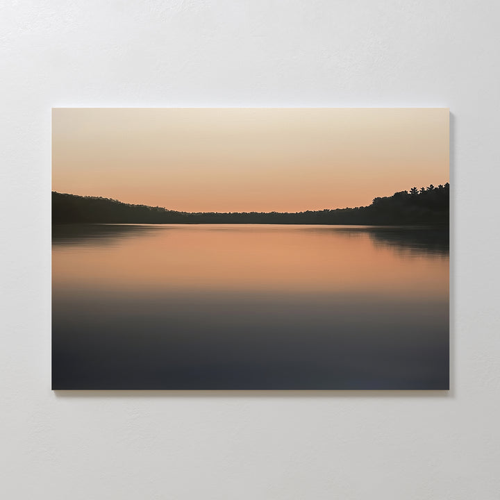 The Still Waters Canvas Art – Horizontal features a minimalist photograph of a calm lake at sunset, with smooth water reflecting orange hues and silhouetted trees for peaceful, serene bedroom decor.