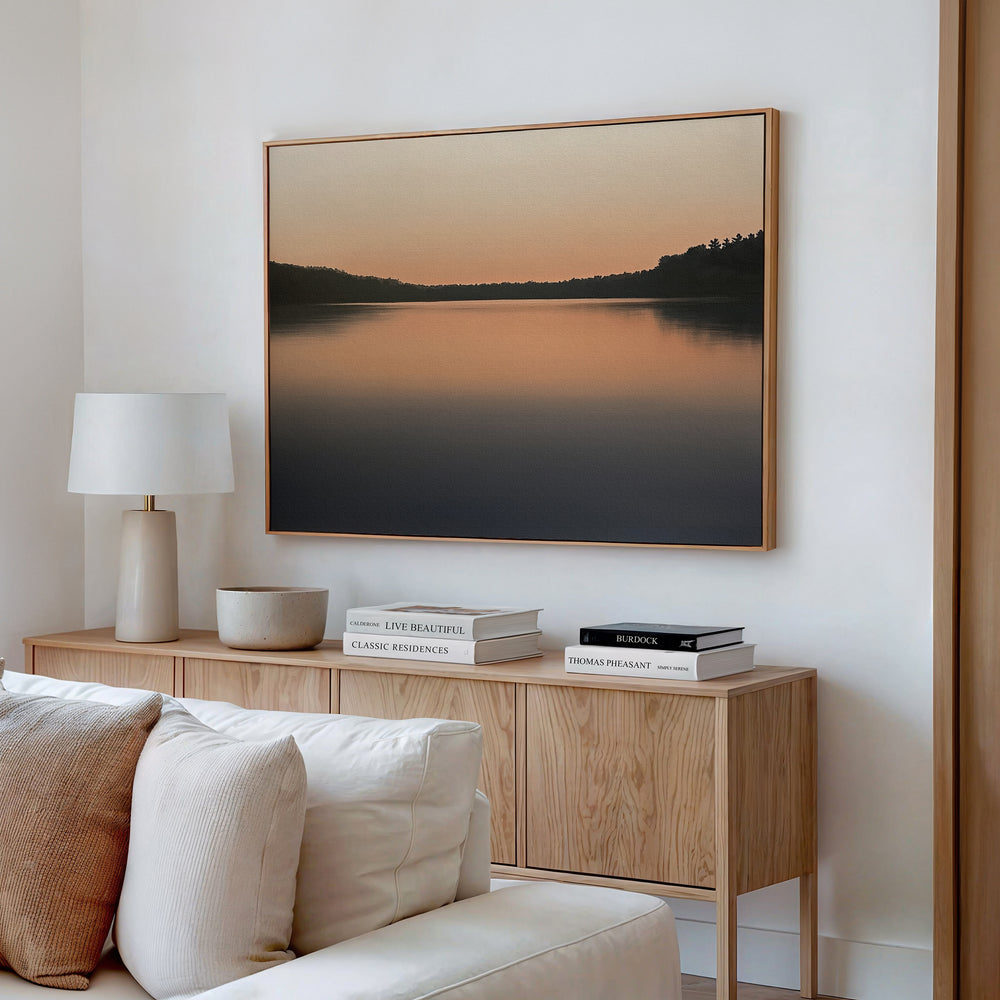 A modern living room features the Still Waters Canvas Art – Horizontal above a wooden sideboard with books, a lamp, and a bowl. A white sofa sits in front, complementing the minimalist and neutral-toned decor.