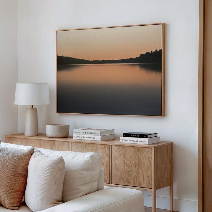 A modern living room features the Still Waters Canvas Art – Horizontal above a wooden sideboard with books, a lamp, and a bowl. A white sofa sits in front, complementing the minimalist and neutral-toned decor.