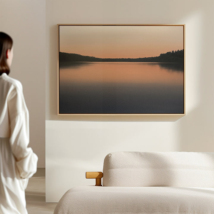 A person in white stands near a bed, admiring the Still Waters Canvas Art – Horizontal, a tranquil print of a serene lake at sunset with soft orange and blue hues—ideal for peaceful bedroom decor.