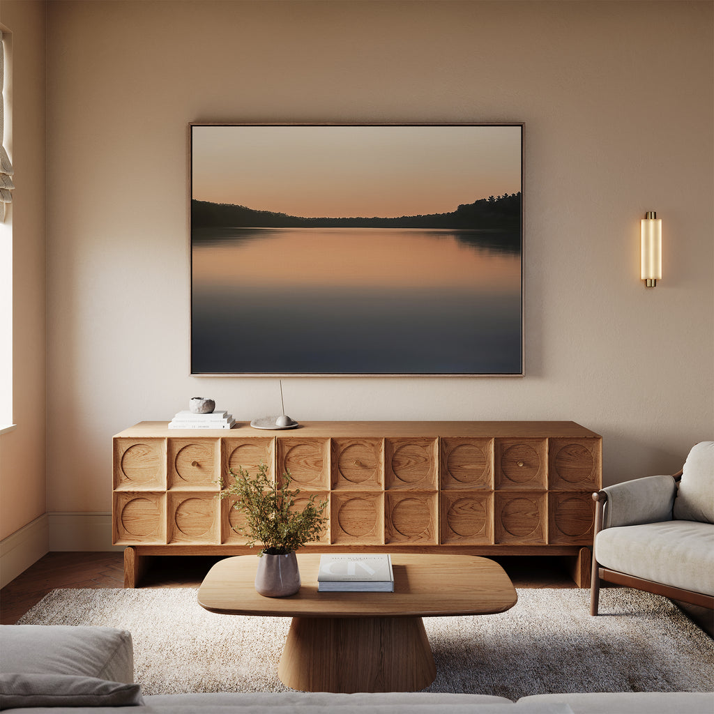 A cozy living room with neutral tones features a wooden sideboard, low coffee table, and soft rug. Above the sideboard hangs the Still Waters Canvas Art – Horizontal, a framed landscape of a serene lake at sunset.