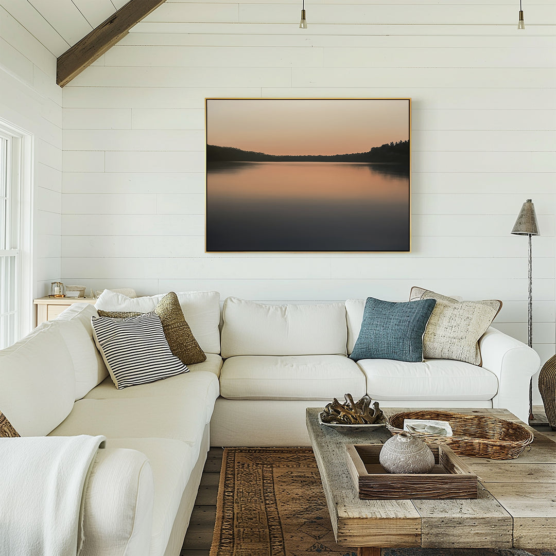 A cozy living room with a white sectional sofa, assorted pillows, rustic wooden coffee table, and the Still Waters Canvas Art – Horizontal depicting a calm lake at sunset on a white shiplap wall.