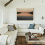 A cozy living room with a white sectional sofa, assorted pillows, rustic wooden coffee table, and the Still Waters Canvas Art – Horizontal depicting a calm lake at sunset on a white shiplap wall.