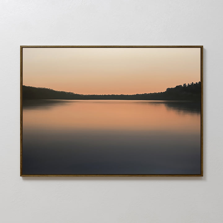 The Still Waters Canvas Art – Horizontal features a tranquil sunset lake scene in soft orange and peach tones with glassy water and silhouetted trees, making it ideal for serene bedroom decor.