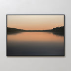 Still Waters Canvas Art – Horizontal: A tranquil canvas print of a calm lake at sunset, with soft orange and peach tones on still waters and dark tree silhouettes—perfect for serene bedroom décor.