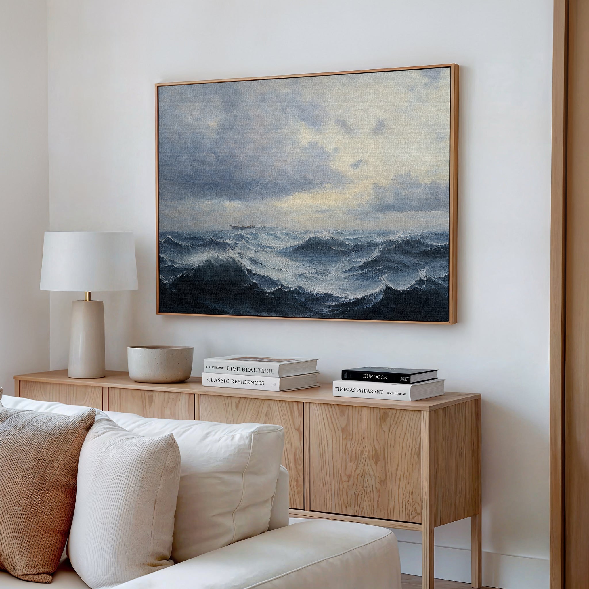 A living room features a light wooden sideboard with stacked books, a white lamp, and a bowl. Above, the Stormy Seas Canvas Art depicting turbulent oceans and cloudy skies hangs on the wall. A cream sofa sits in front.