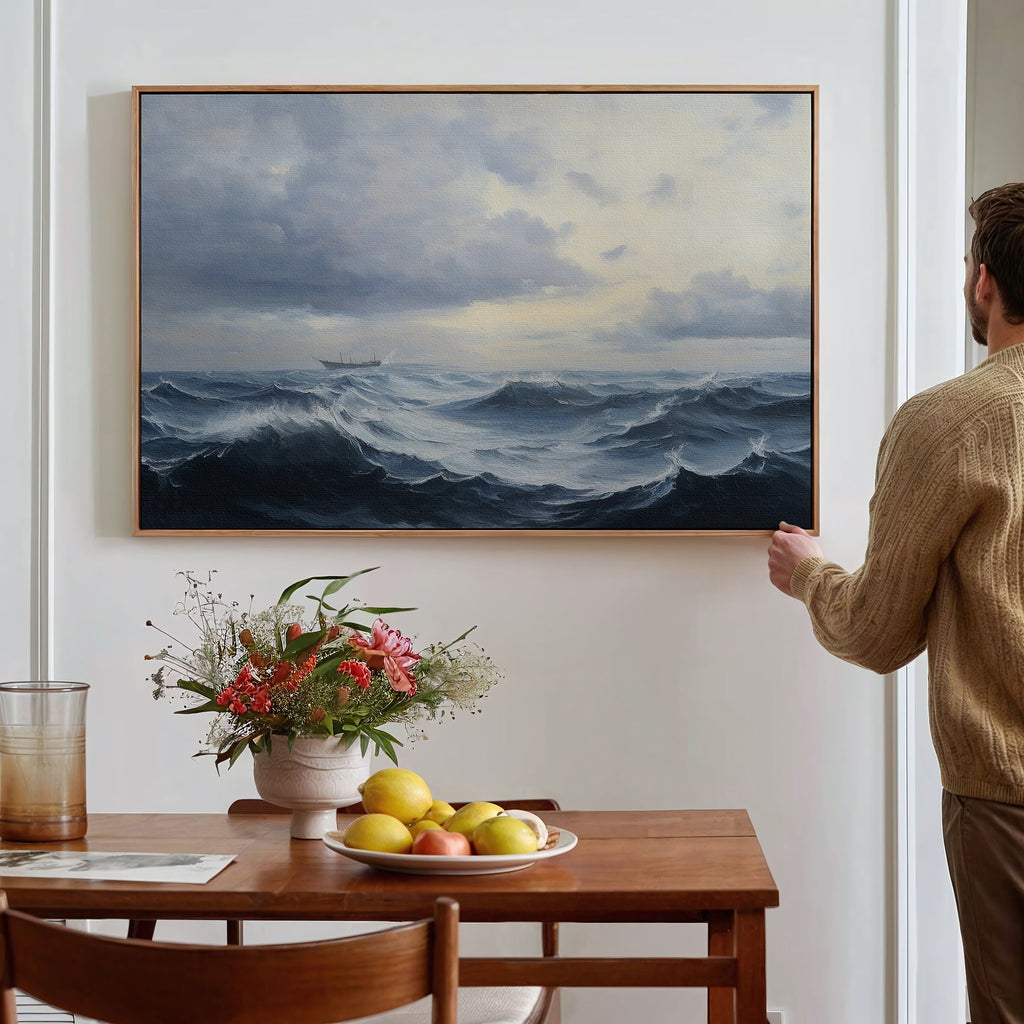 A person hangs a Stormy Seas Canvas Art above a wooden dining table, which holds a bowl of fruit, a vase of flowers, and a glass of water with a book beside it.