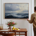 A person hangs a Stormy Seas Canvas Art above a wooden dining table, which holds a bowl of fruit, a vase of flowers, and a glass of water with a book beside it.