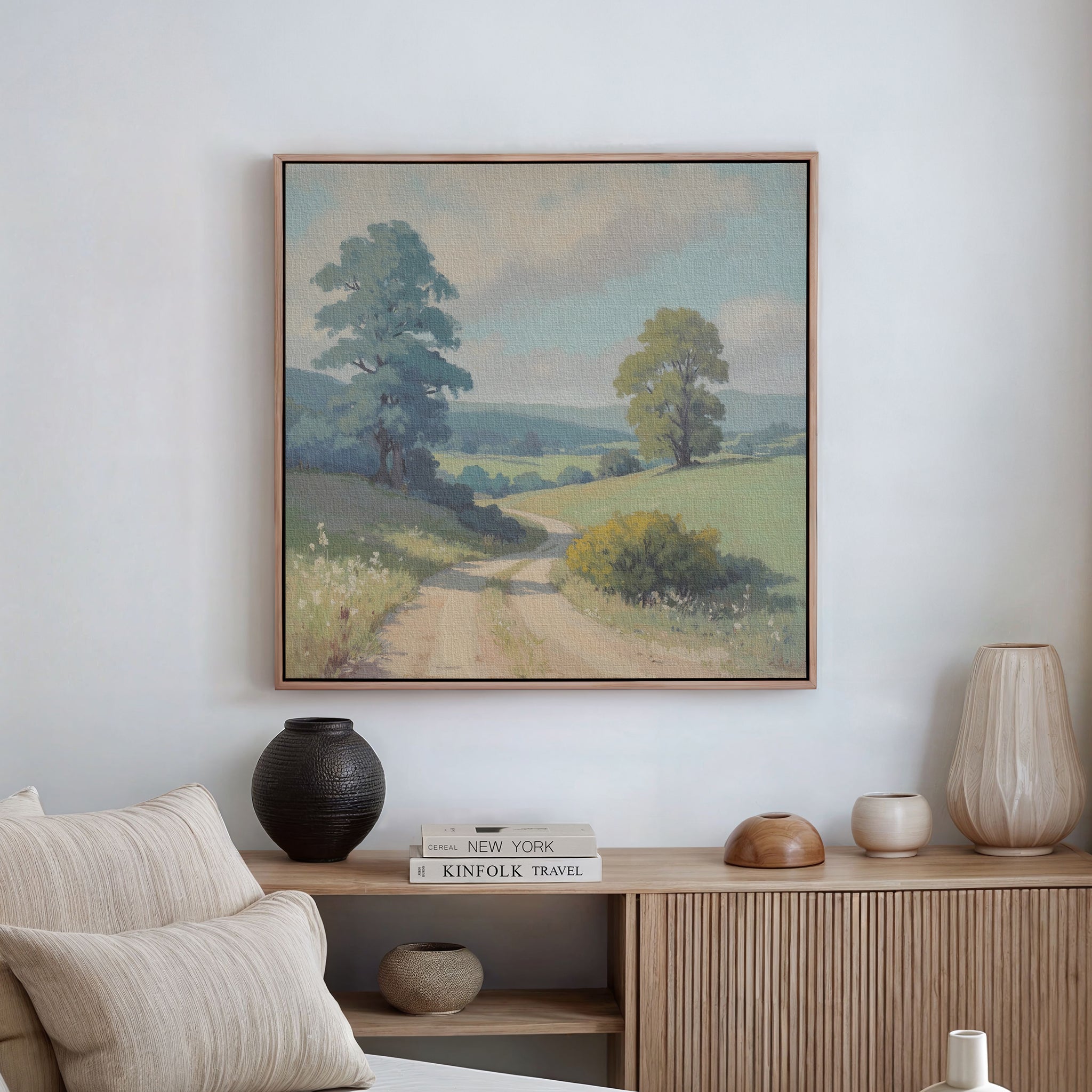 The Summer Dale Canvas Art, depicting a dirt road through green fields and trees, hangs above a wooden sideboard with vases, a bowl, and books below. A light-colored sofa is partially visible, enhancing the rustic charm of the space.
