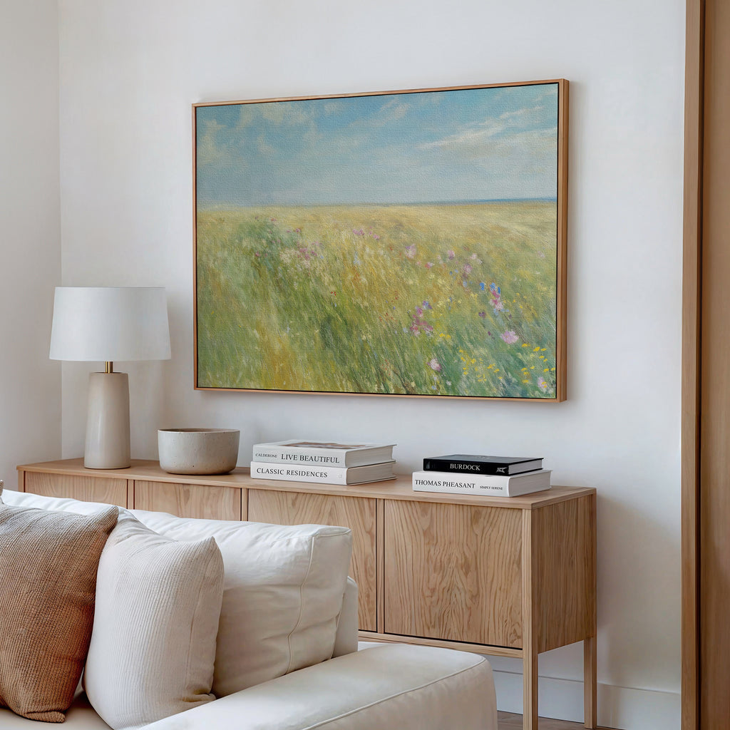 A cozy living room features a wooden sideboard with books, a lamp, and a bowl. Above hangs the Summers Embrace Canvas Art depicting a bright field and blue sky. A cream sofa in the foreground enhances the inviting atmosphere.