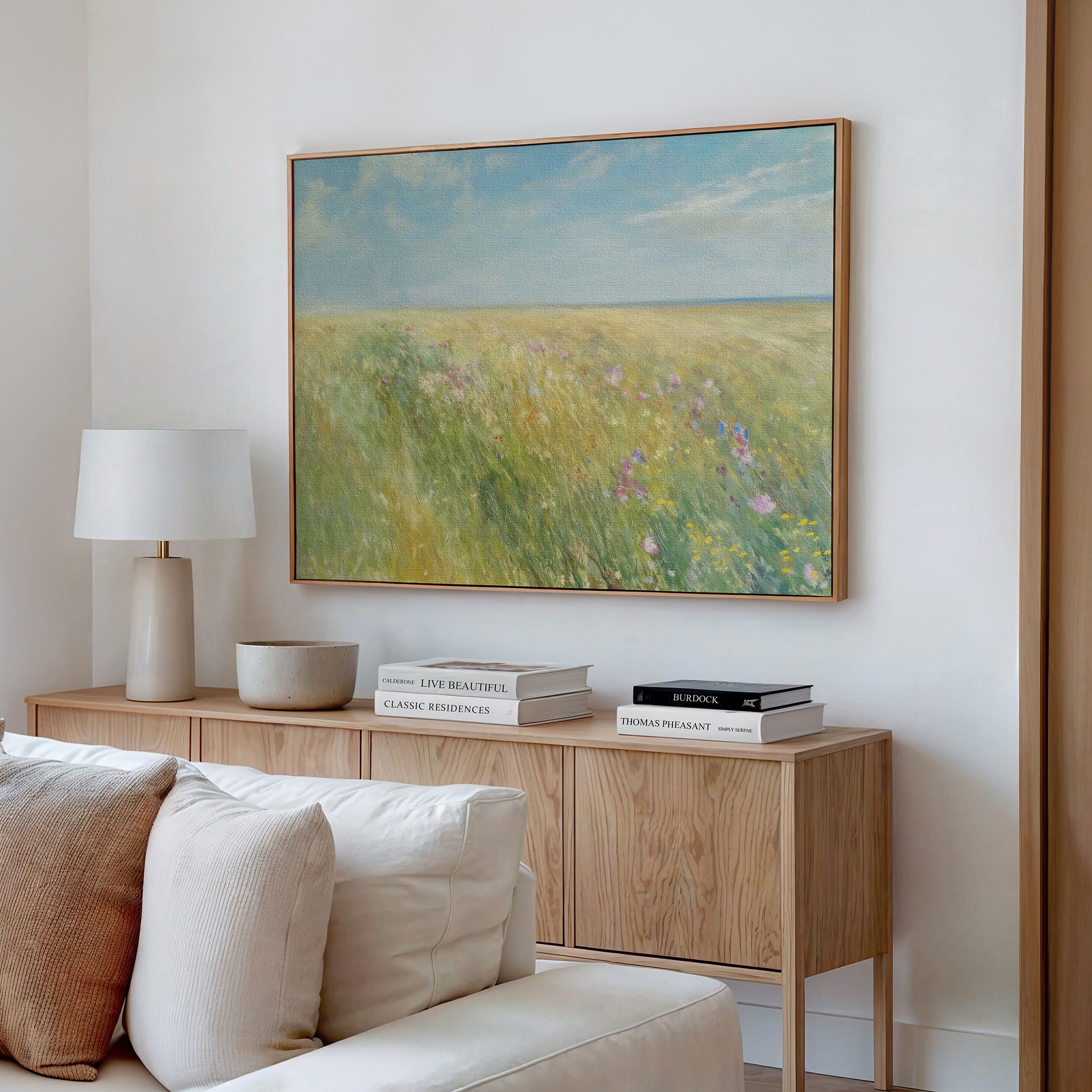A cozy living room features a wooden sideboard with books, a lamp, and a bowl. Above hangs the Summers Embrace Canvas Art depicting a bright field and blue sky. A cream sofa in the foreground enhances the inviting atmosphere.