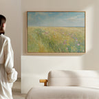A woman stands in a bright, minimalist room, admiring the Summers Embrace Canvas Art—a landscape print of a grassy field with wildflowers and blue sky—hung on a light wall above a beige sofa.