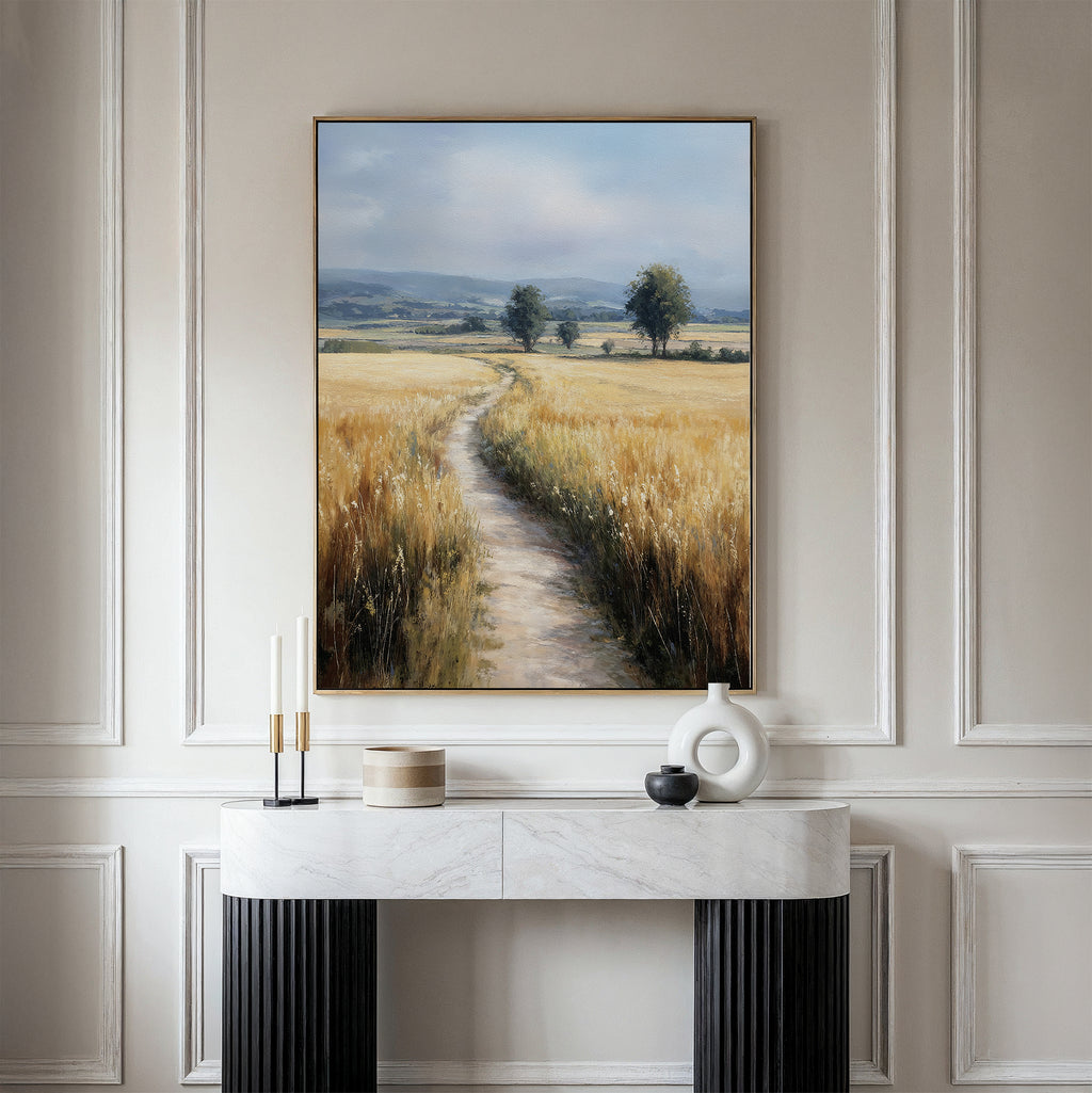 A marble console table with modern decor sits against a white paneled wall. Above it hangs the Sunlit Passage Canvas Art, depicting a country path winding through golden fields toward distant trees and hills under a blue sky.