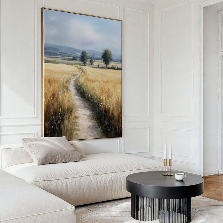 A modern living room featuring a beige sectional sofa, a round black coffee table with candles and a cup, and the Sunlit Passage Canvas Art—a nature-inspired country path scene—displayed above.