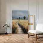 A room with herringbone wood floors, white walls, a white cushioned chair, a small black round table with candles and books, and the Sunlit Passage Canvas Art of a path through a golden field leaning against the wall.