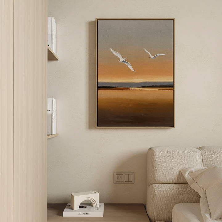 The Sunset Flight Canvas Art, featuring two white birds soaring over a serene sunset landscape, is displayed above a light sofa while books and a decorative object rest on the adjacent shelf.