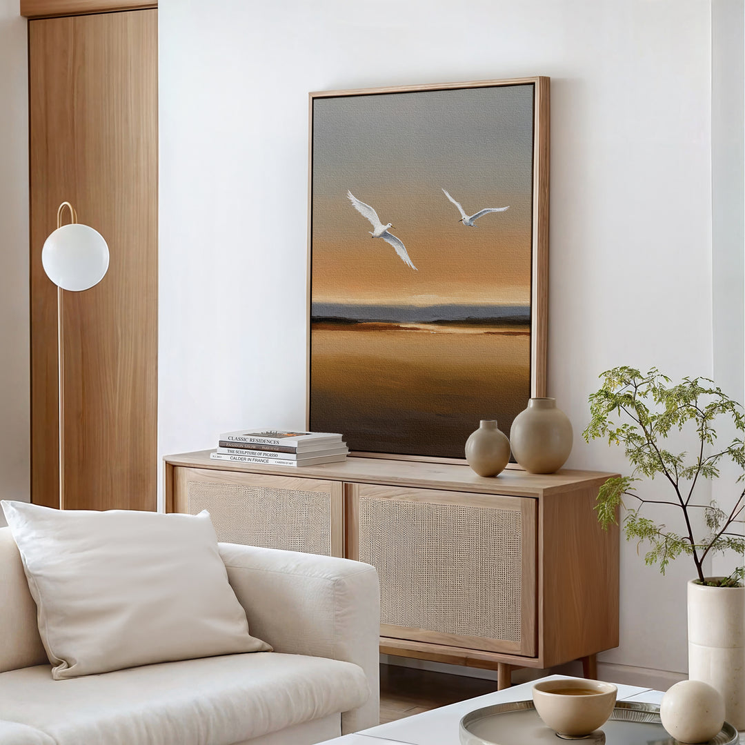 A minimalist living room with a light wood sideboard, neutral decor, the Sunset Flight Canvas Art showing two birds at sunset, a white sofa with cushions, potted plant, and a round light fixture.