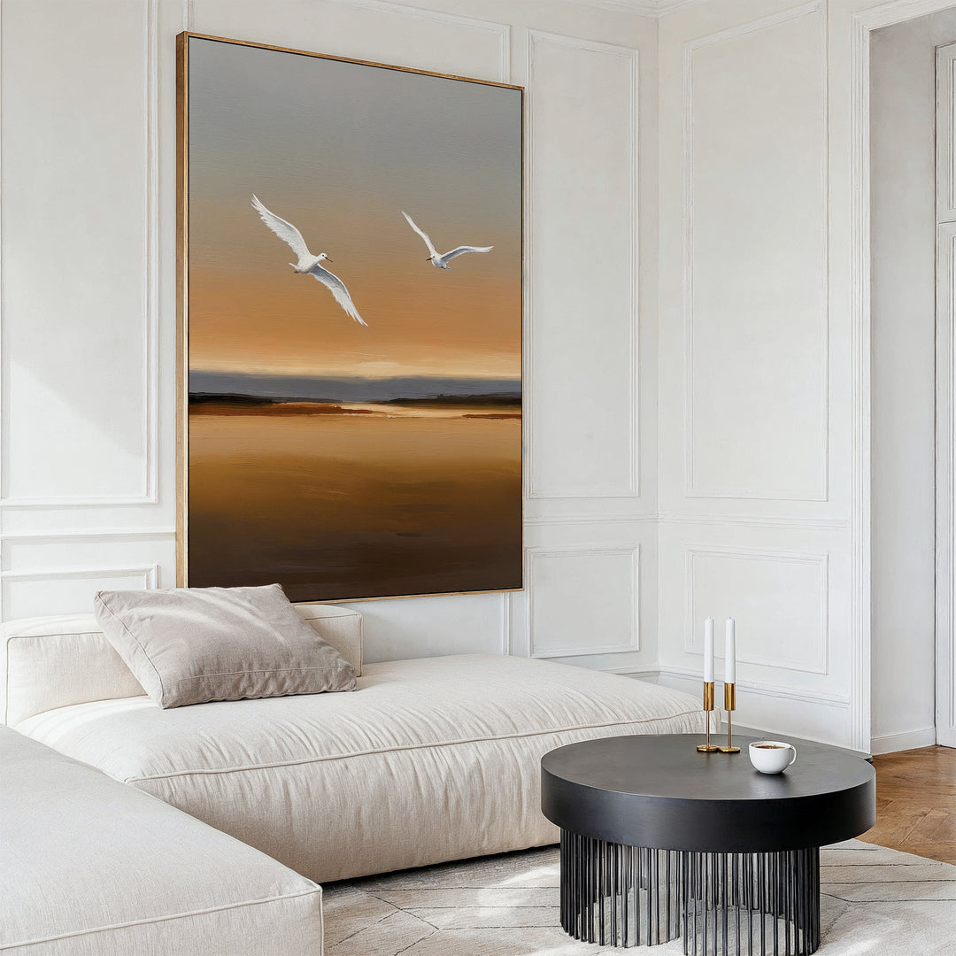 A modern living room with a cream sofa, a round black coffee table holding two candlesticks and a cup, and the striking Sunset Flight Canvas Art featuring two white birds flying over a golden landscape on the wall.