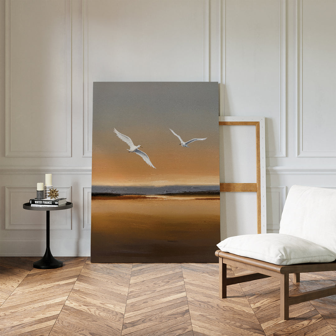 A minimalist room showcases the Sunset Flight Canvas Art—depicting two white birds over a sunset beach—leaning against a white paneled wall, complemented by a cushioned chair and a small round table with candles and books.