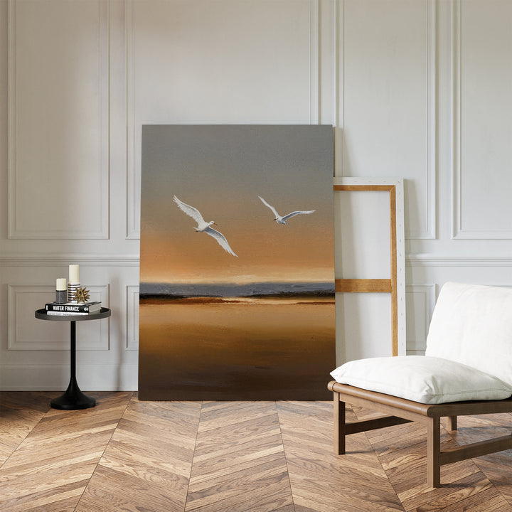 A minimalist room showcases the Sunset Flight Canvas Art—depicting two white birds over a sunset beach—leaning against a white paneled wall, complemented by a cushioned chair and a small round table with candles and books.