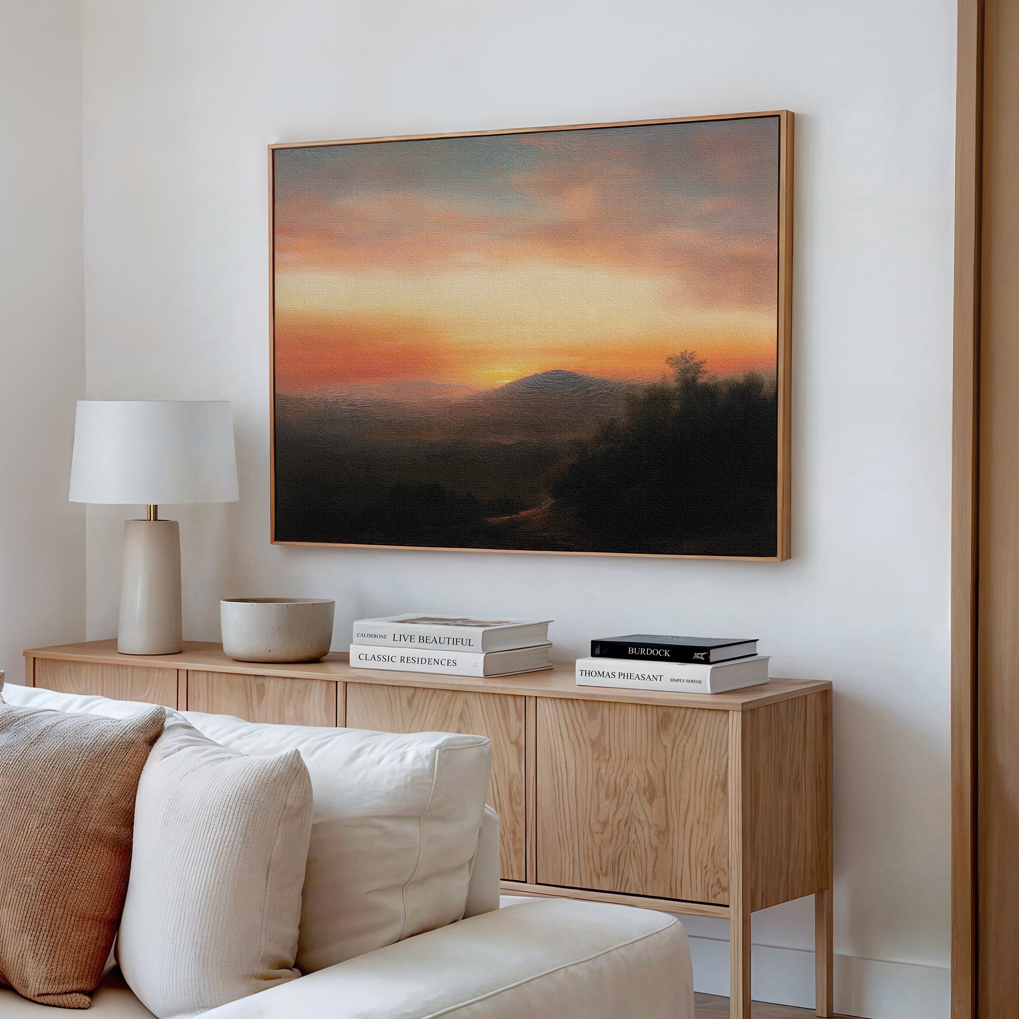 A cozy living room with a wooden sideboard, neutral decor, a lamp, and books. Above the sideboard hangs Sunset Reflection Canvas Art—a vibrant framed landscape of a colorful sunset over hills. A white sofa is partly visible in front.