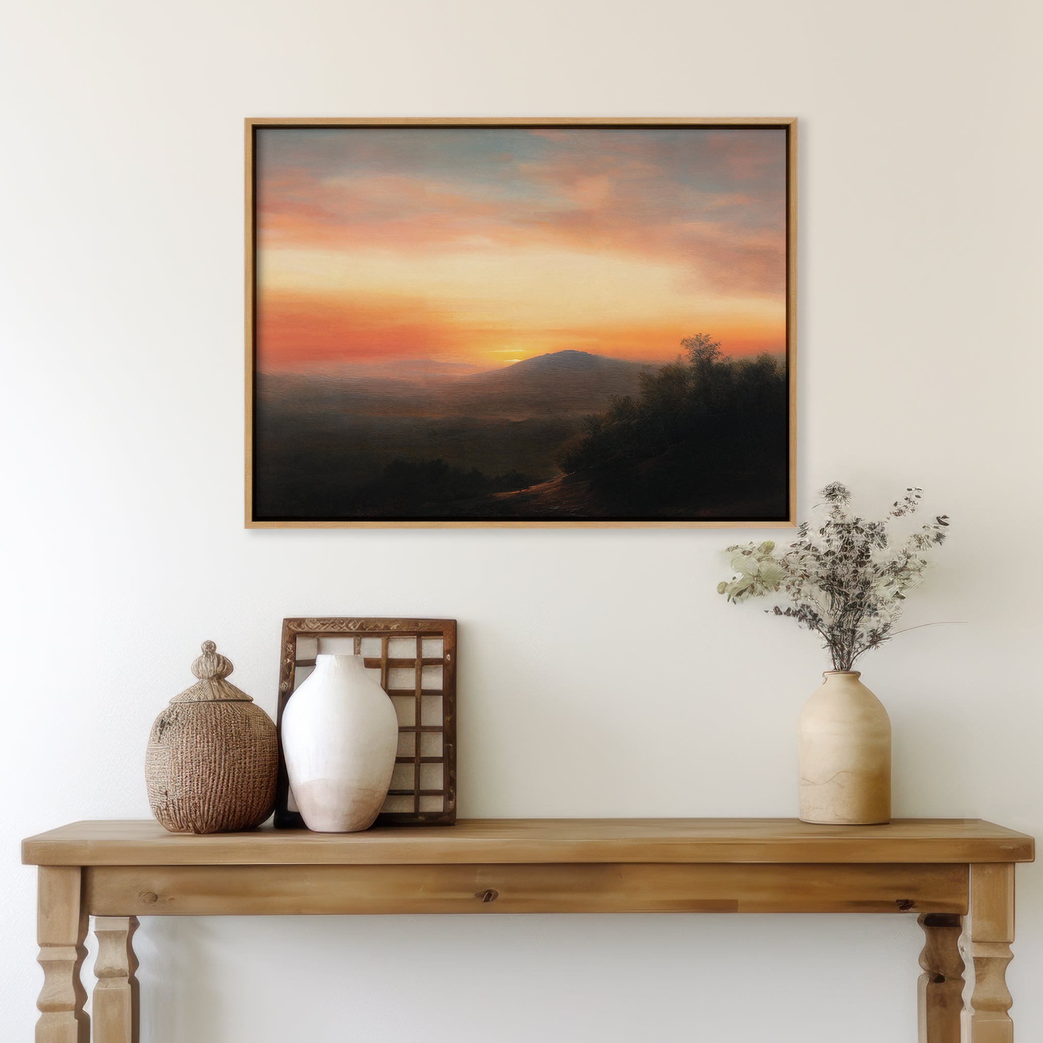 A wooden table with two vases, a decorative basket, and a small wooden frame stands against a light wall, above which hangs the Sunset Reflection Canvas Art depicting a colorful sunset over rolling hills.