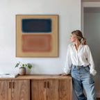 A woman in a white blouse and jeans stands by a wooden sideboard, admiring the Sunset Vapor Canvas Art with blue and brown shapes above her. A potted plant and books add cozy touches to the modern interior.