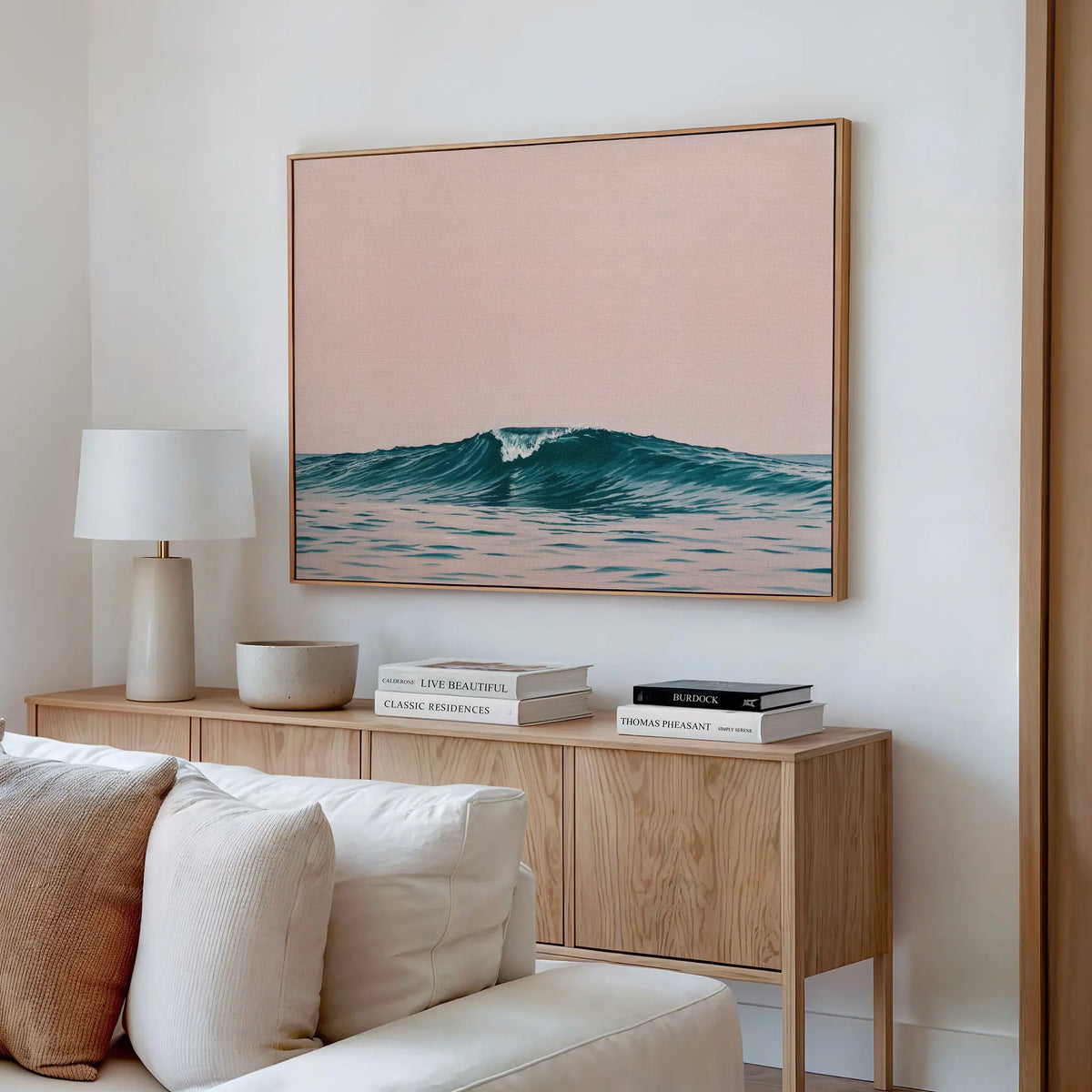 A modern living room features a light wooden sideboard, white lamp, books, and a beige sofa. Above the sideboard hangs the Sunset Wave Canvas Art—a large pastel wave print set against a soft pink sky.
