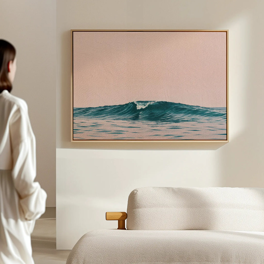 A person stands in a minimal, light-filled room beside a cream-colored sofa, admiring the Sunset Wave Canvas Art—a framed ocean wave beneath a soft pink sky—serving as beach-inspired wall decor.