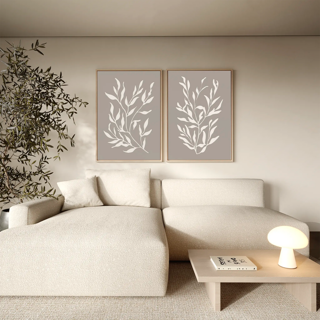 A modern living room featuring a cream sectional sofa, a small wooden coffee table with a lamp and books, a potted plant, and the Taupe Botanical Set of 2 wall art prints for contemporary interiors.