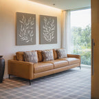 A modern living room featuring a tan leather sofa with four patterned cushions, contemporary decor, a Taupe Botanical Set of 2 prints on the wall, a decorative vase, and a window with sheer curtains overlooking greenery.