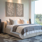 A modern bedroom with a large bed, white and gray bedding, four white pillows, the Taupe Botanical Set of 2 above the headboard, large floor vases, and a floor-to-ceiling window showcasing outdoor greenery.
