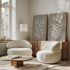 A minimalist living room features two rounded white armchairs, a low wooden coffee table, and the Taupe Botanical Set of 2 prints on a sideboard. Large windows fill the space with natural light for a cozy, modern vibe.