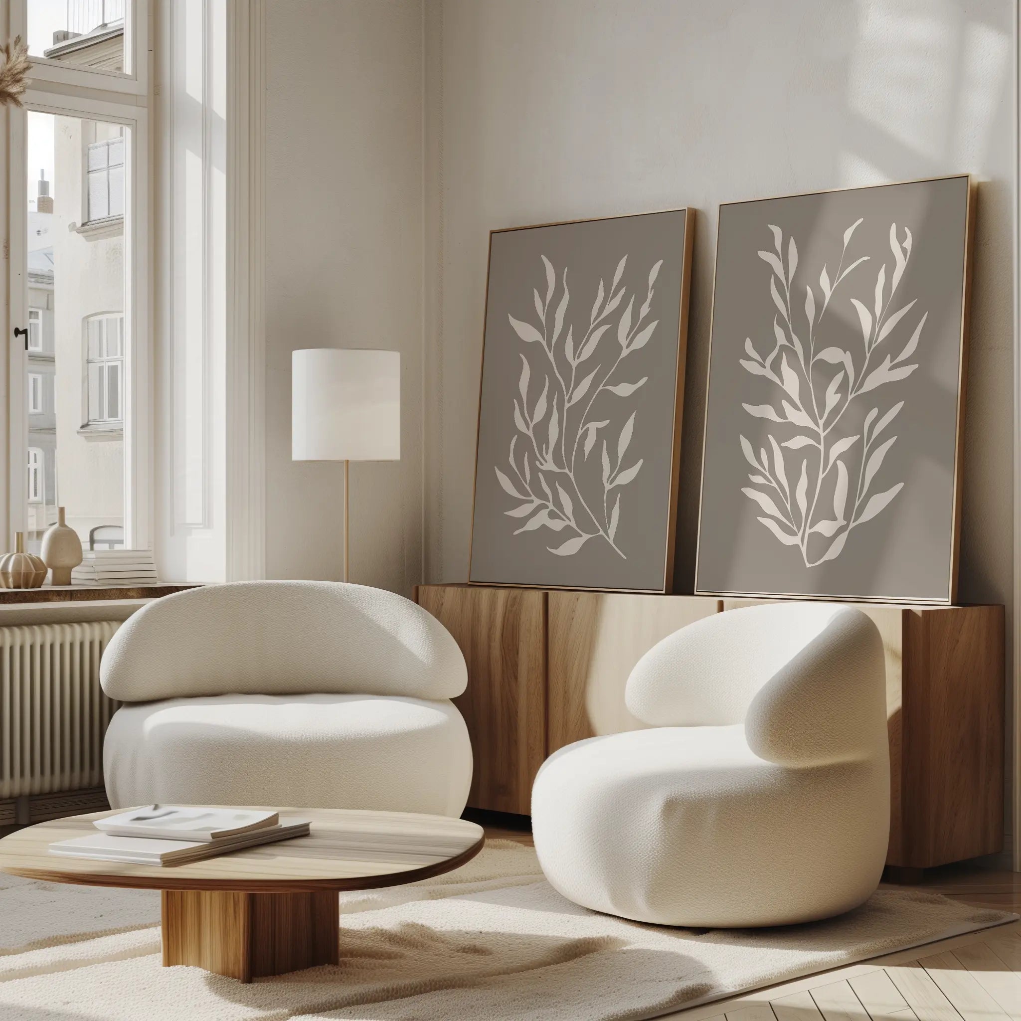 A minimalist living room features two rounded white armchairs, a low wooden coffee table, and the Taupe Botanical Set of 2 prints on a sideboard. Large windows fill the space with natural light for a cozy, modern vibe.