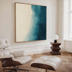 A modern living room with a white leather lounge chair and ottoman, wooden side table with a white lamp, Teal Tide Canvas Art in blue and cream tones on the wall, light wood herringbone floor, and a large window.